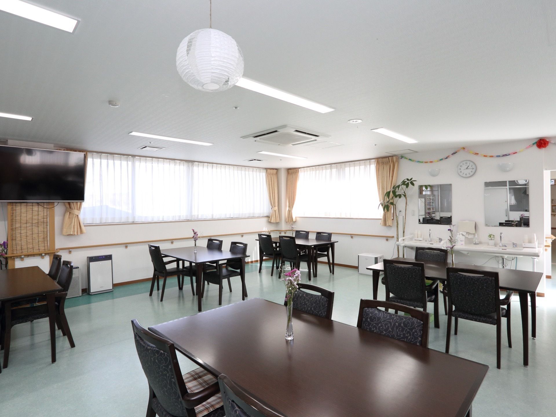 多目的室には焦げ茶色のテーブルと椅子が整然と配置されており、入居者様の通行の際に障害にならないように考慮されている。