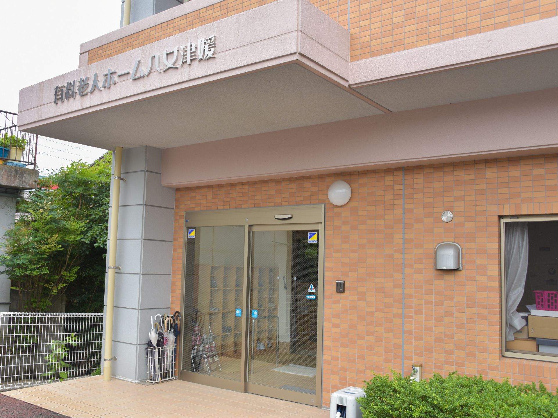 玄関はガラス戸の自動ドア。脇に傘立てが置いてある。玄関の上部には「有料老人ホーム八女津媛」の名前が掲げられている。
