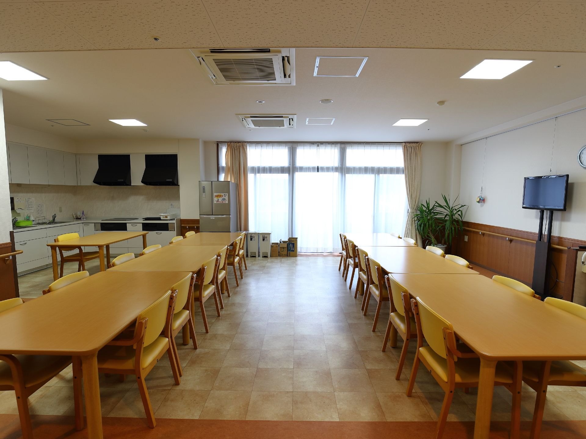ダイニングの壁面の高い位置には大きな液晶テレビが設置されている。部屋の隅には観葉植物の鉢植えが置かれている。