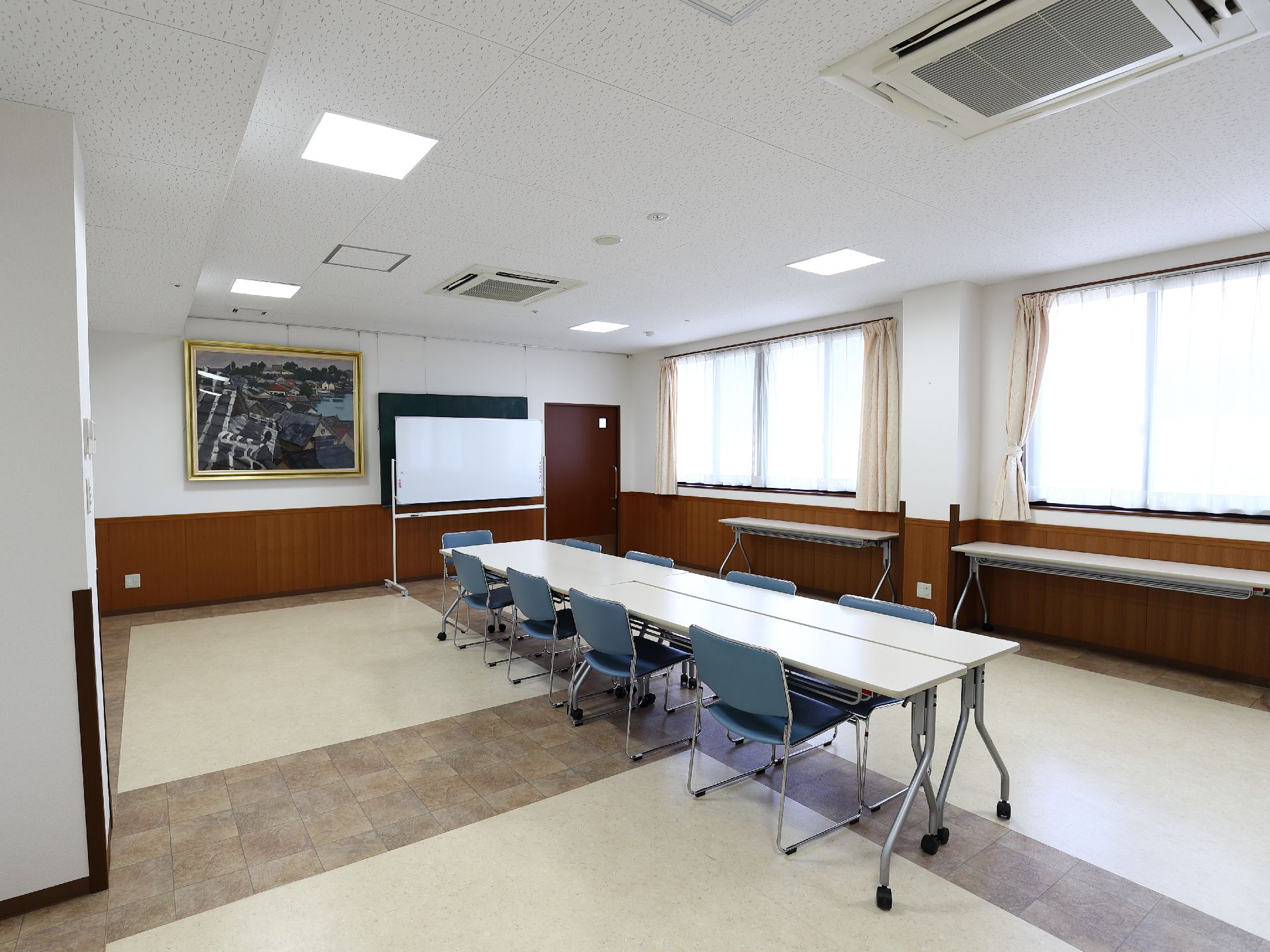 窓が大きくて開放的な広々とした会議室。長い会議用机に9人分の椅子、可動式のホワイトボードが置いてある。