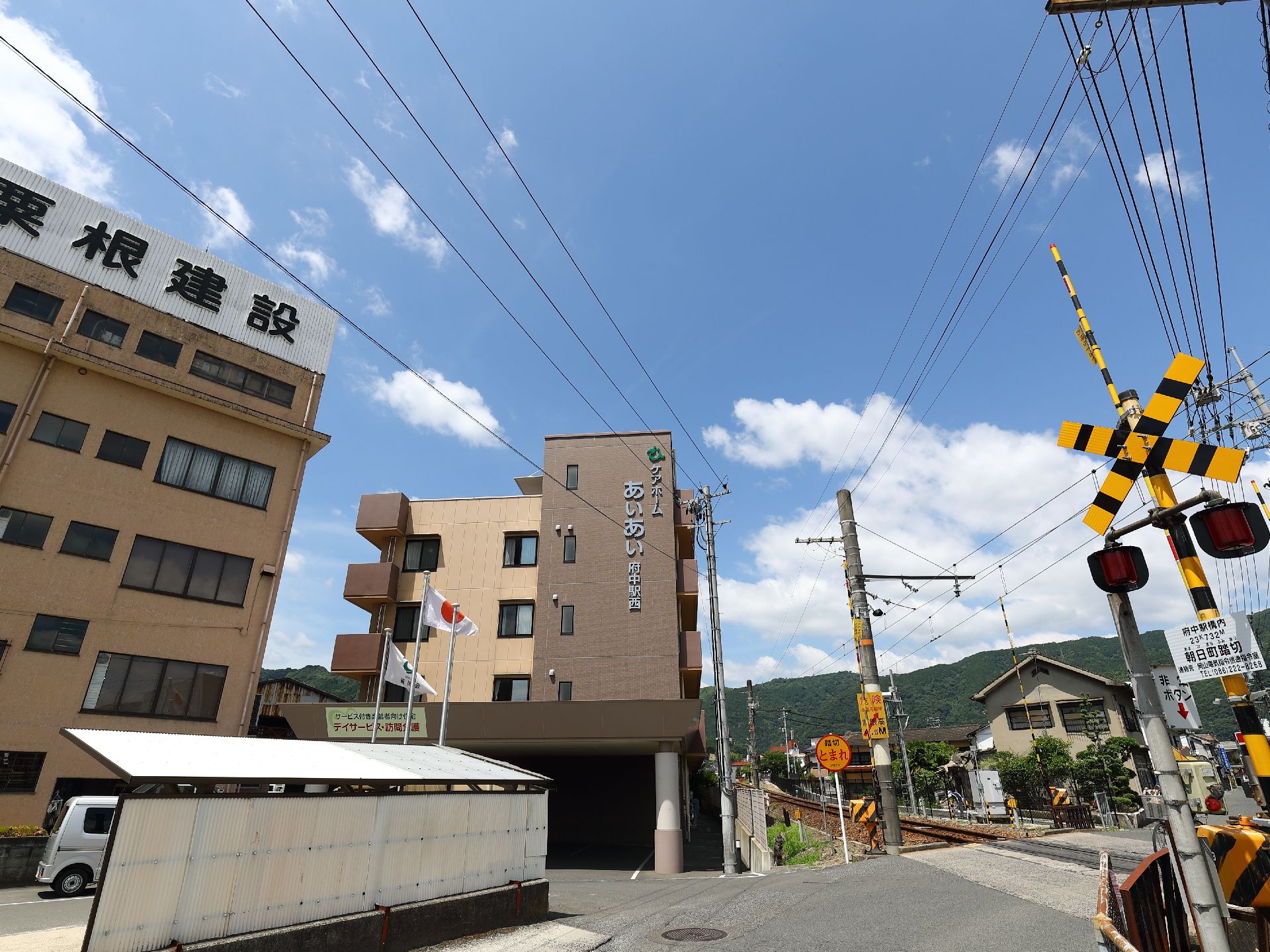 4階建ての建物で、踏切のすぐ横に建っている。周辺には高い建物は少なく、背後には青空と山並みが見える。