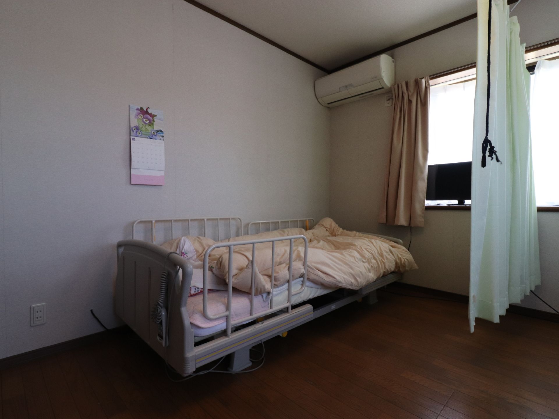 2人用の居室の壁際に置かれた介護用ベッドである。壁にはカレンダーが掛けてあり、天上にはエアコンが設置されている。