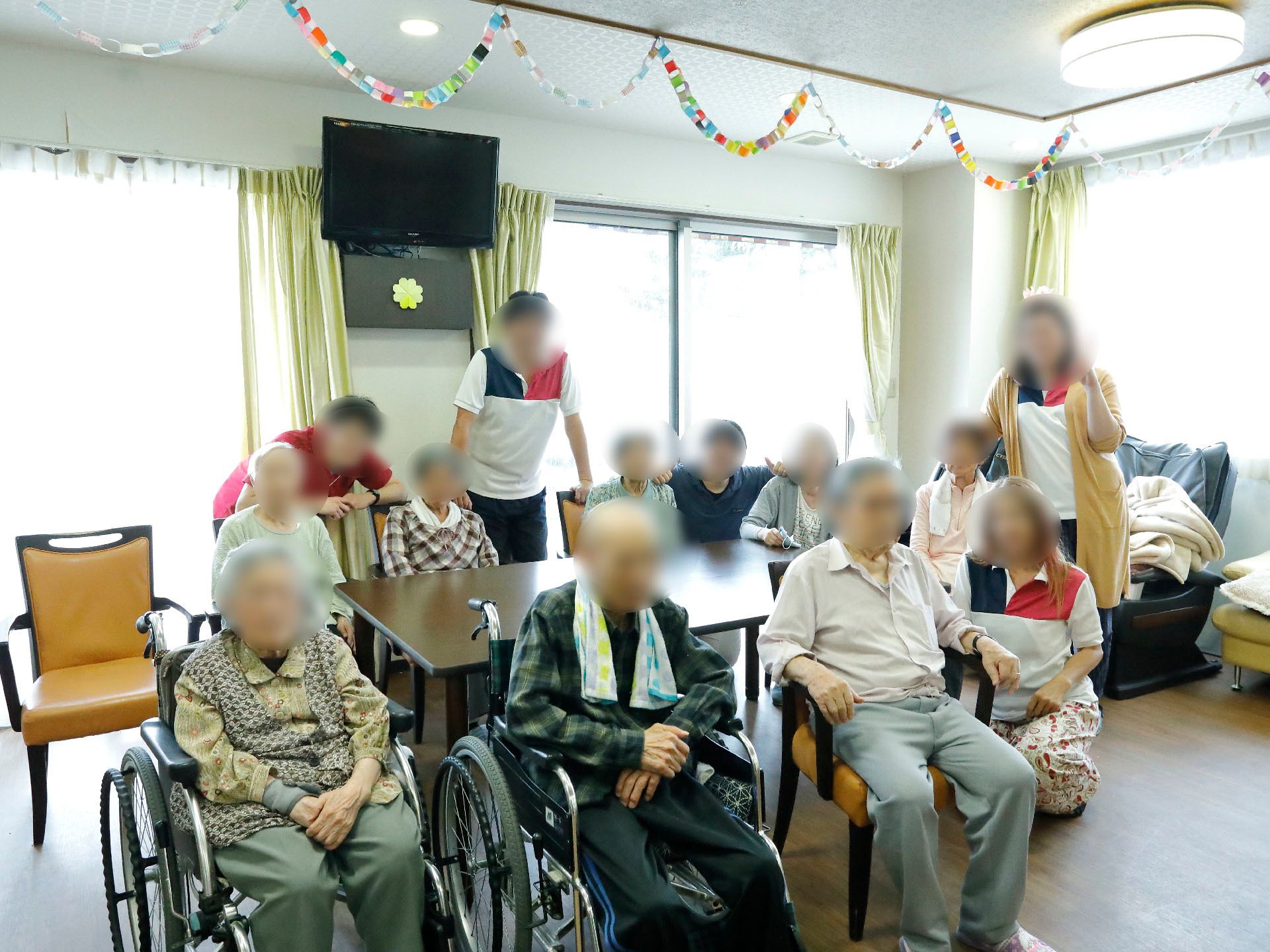 施設の利用者とスタッフが共有スペースに集まっている写真である。スタッフは男性3人と女性2人で、利用者のすぐ横に位置している。