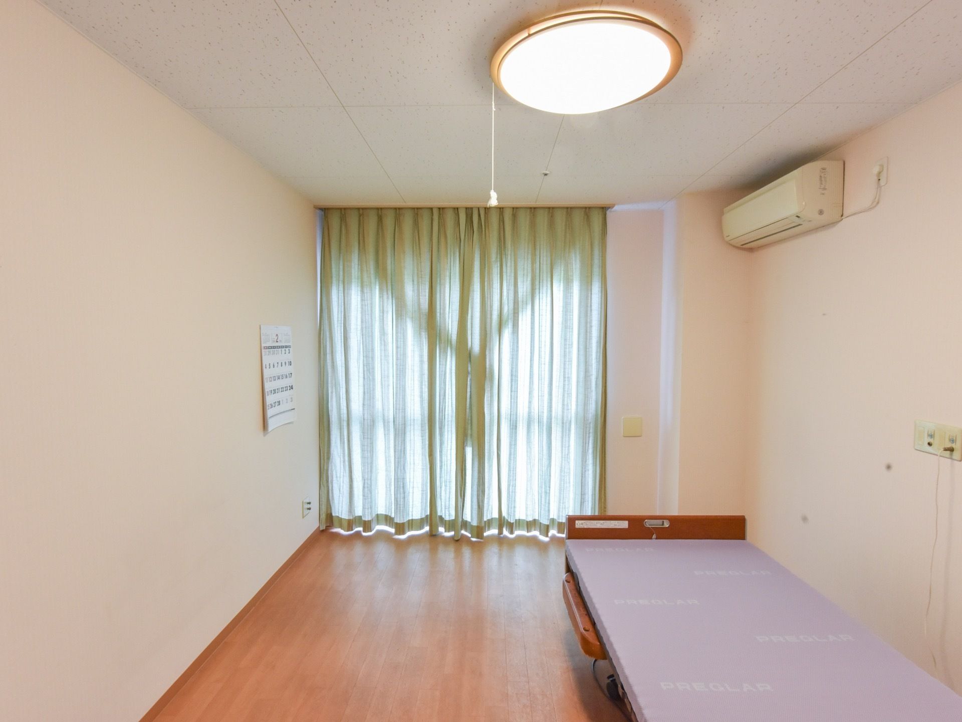 エアコンが完備された居室。大きな窓には緑色のカーテンが取り付けられており、壁にはカレンダーが掛けられている。