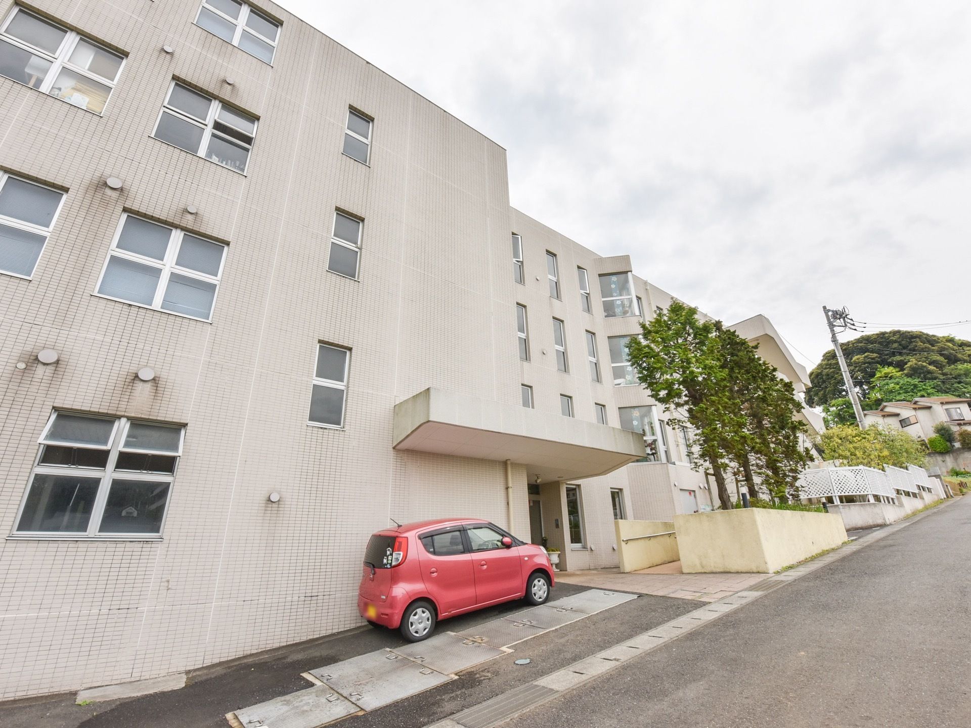 坂の途中に建てられた緑に囲まれているグループホーム。白いタイル調の外壁で、マンションのような４階建ての建物である。