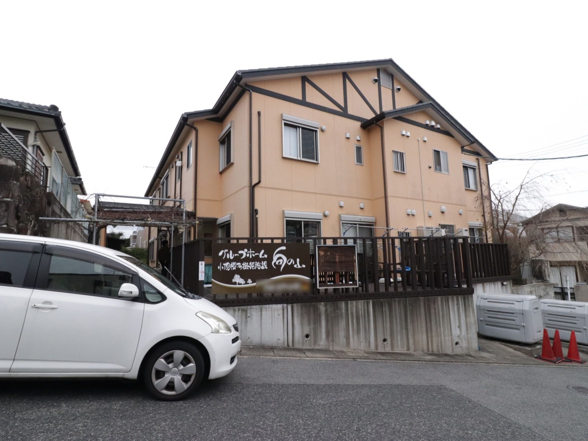 木造２階建ての建物は１階がグループホーム、２階が小規模多機能型居宅介護になっている。道に面した柵には、施設名が書かれた看板が設置されている。