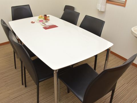 部屋の角部分に白い長方形のテーブルが置かれ、黒い椅子が6脚セットされている。壁際には予備の椅子が1脚置かれている。