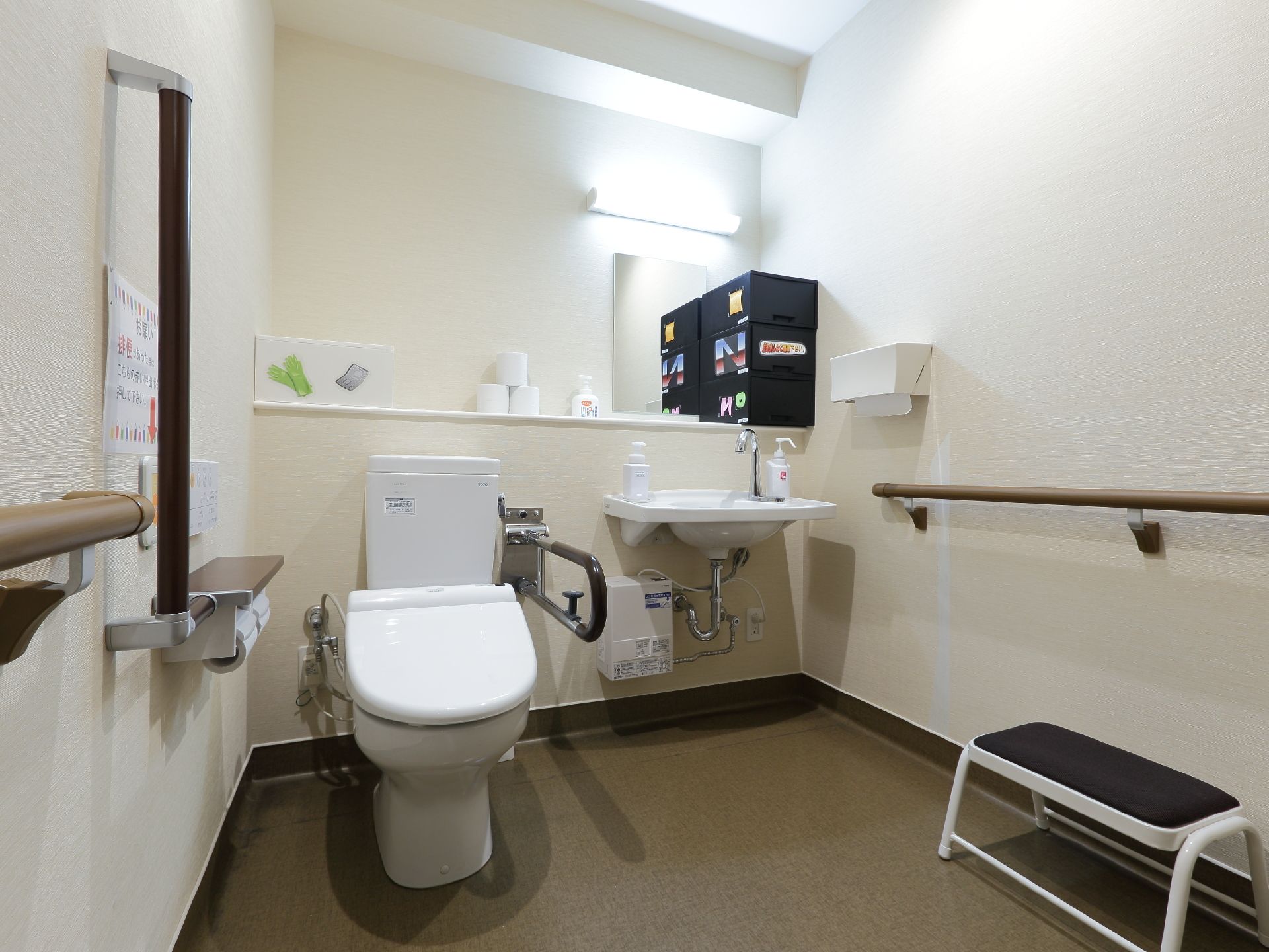 温水洗浄便座付きの洋式トイレが設置されている。壁には手すりやナースコールが付いている。隣には鏡の付いた洗面台が置かれている。