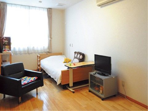 居室の窓際にベッドと小棚を置いている。窓から離れた部屋の側面に、エアコンが設置してある。テレビの前には椅子を用意している。