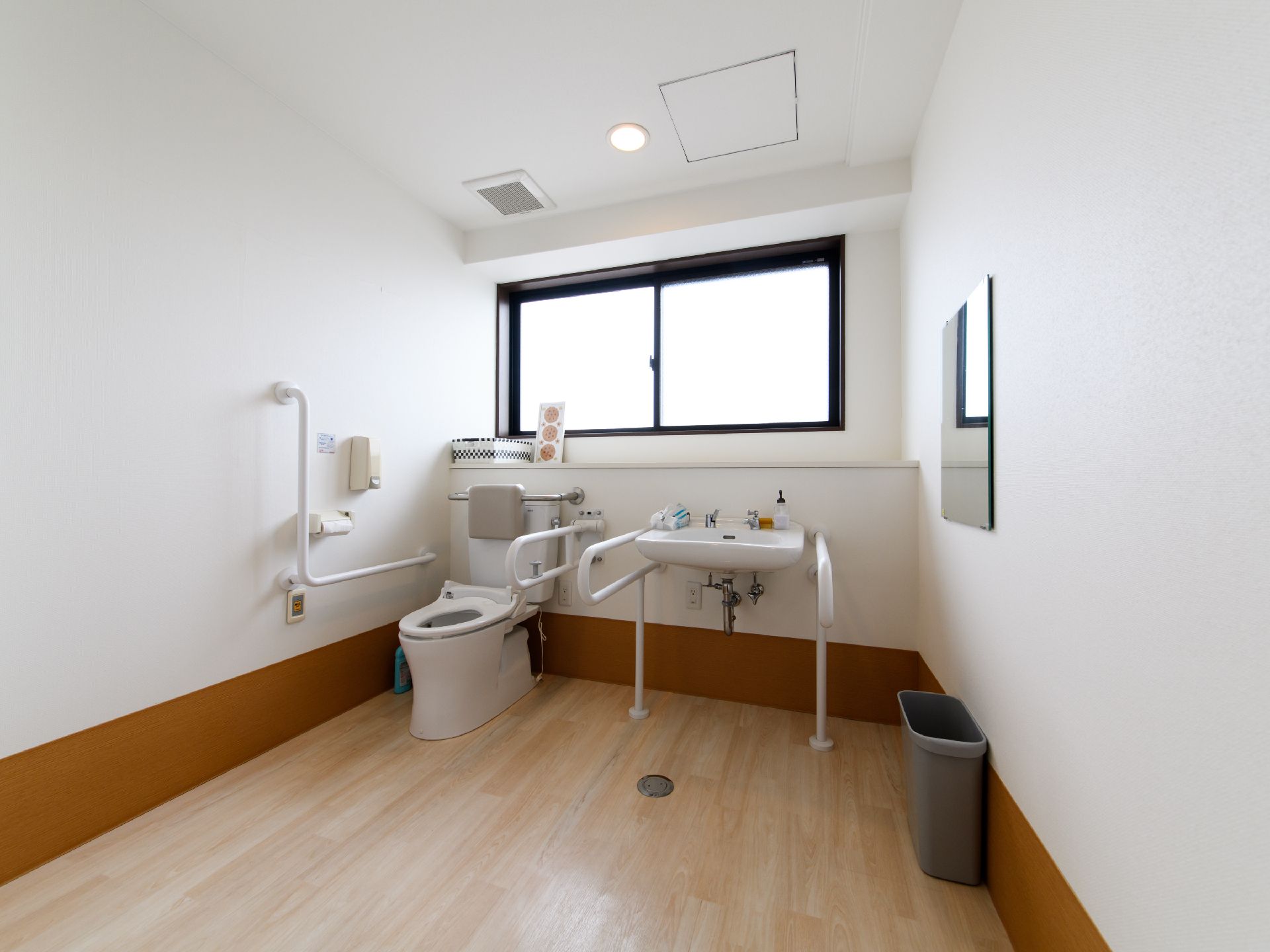 トイレには広い空間があり、正面の壁には窓が設けられている。洋式便器の右隣には洗面台があり、右側の壁には鏡がある。