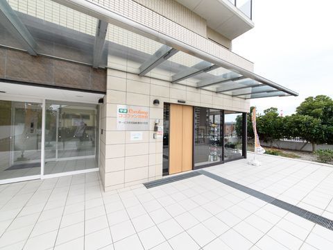 施設の玄関前の床は明るいベージュ色のタイル敷きである。右端にはのぼりが立てられ、上部には大きな透明の屋根がある。