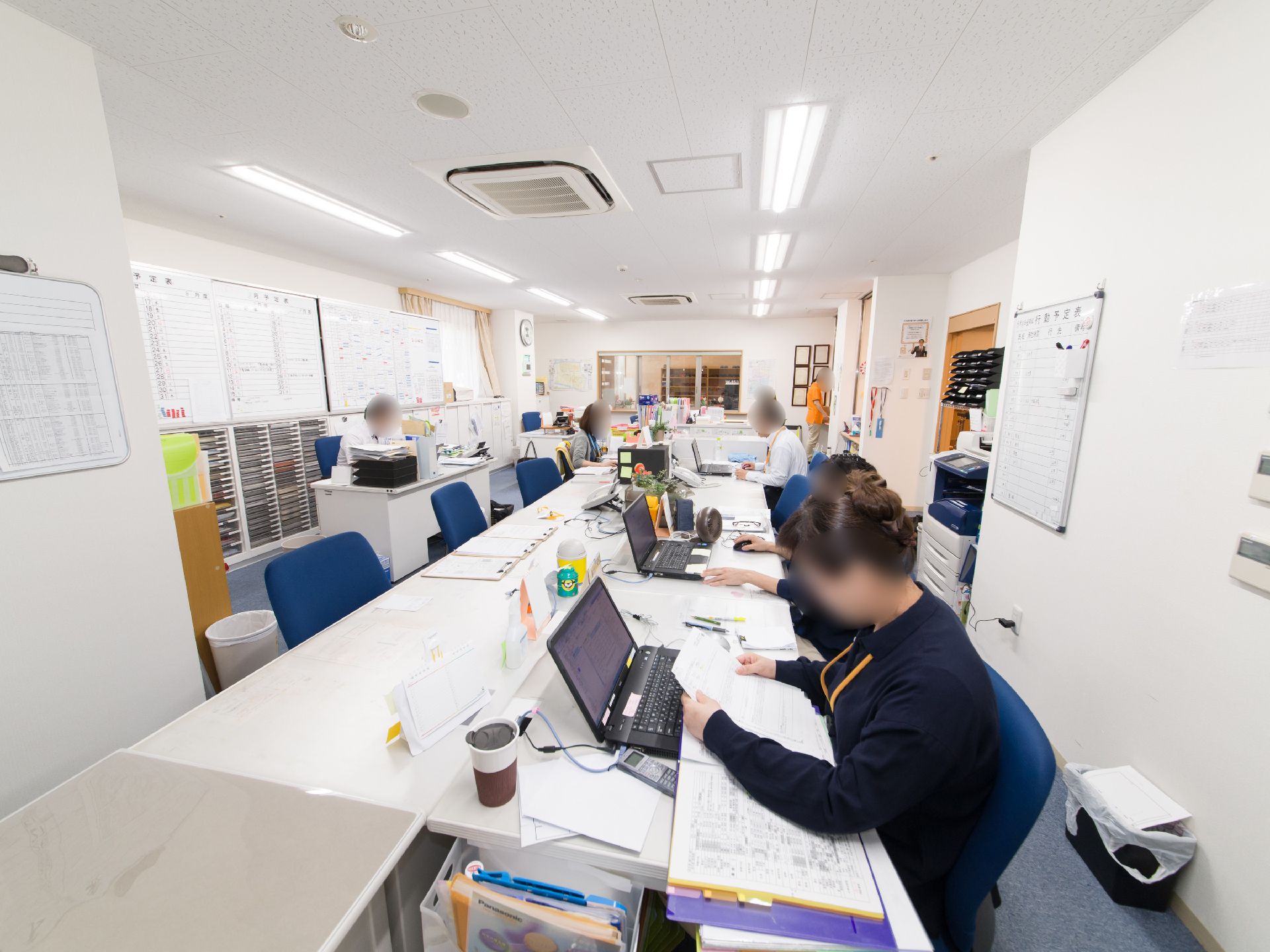 事務室内部には、書類を見たりパソコンを扱ったりしているスタッフがたくさん着席している。壁にはホワイトボードが設置されている。