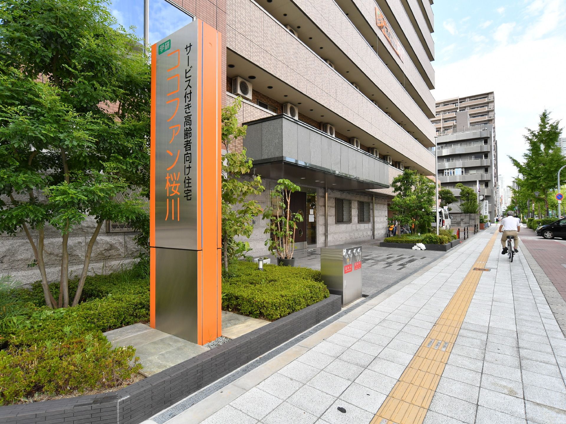 施設外観の手前には、施設名の書かれた看板が掲げられている。施設前は一般道路となっており、自転車で通り過ぎている人がいる。