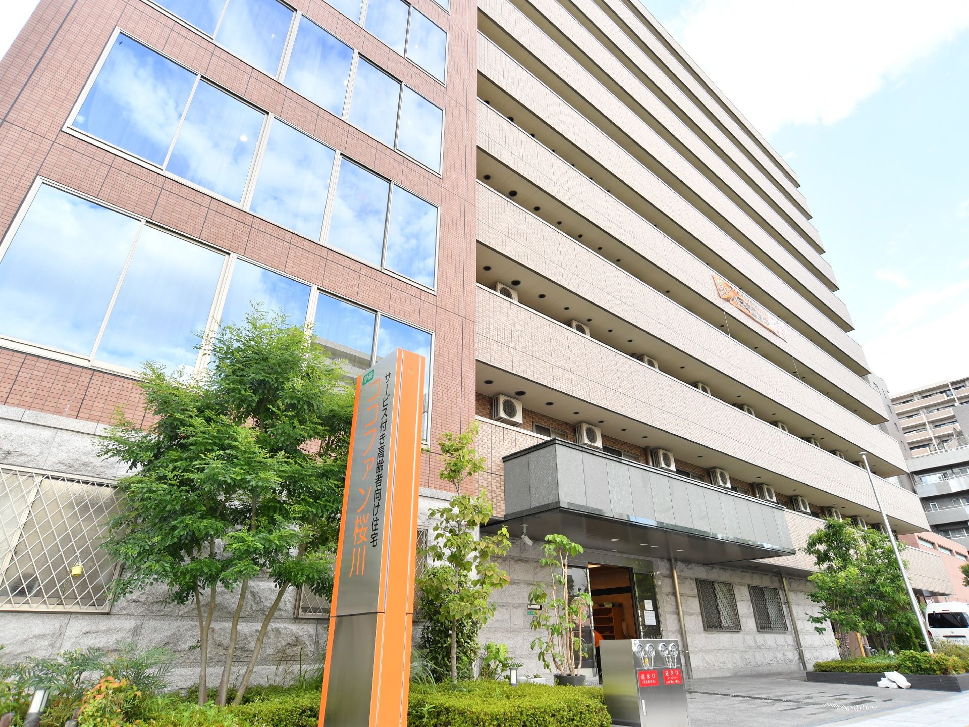 正面に建物がある。左手前には施設名が書かれた長方形型の看板が立っている。その奥には消火設備が設置されている。