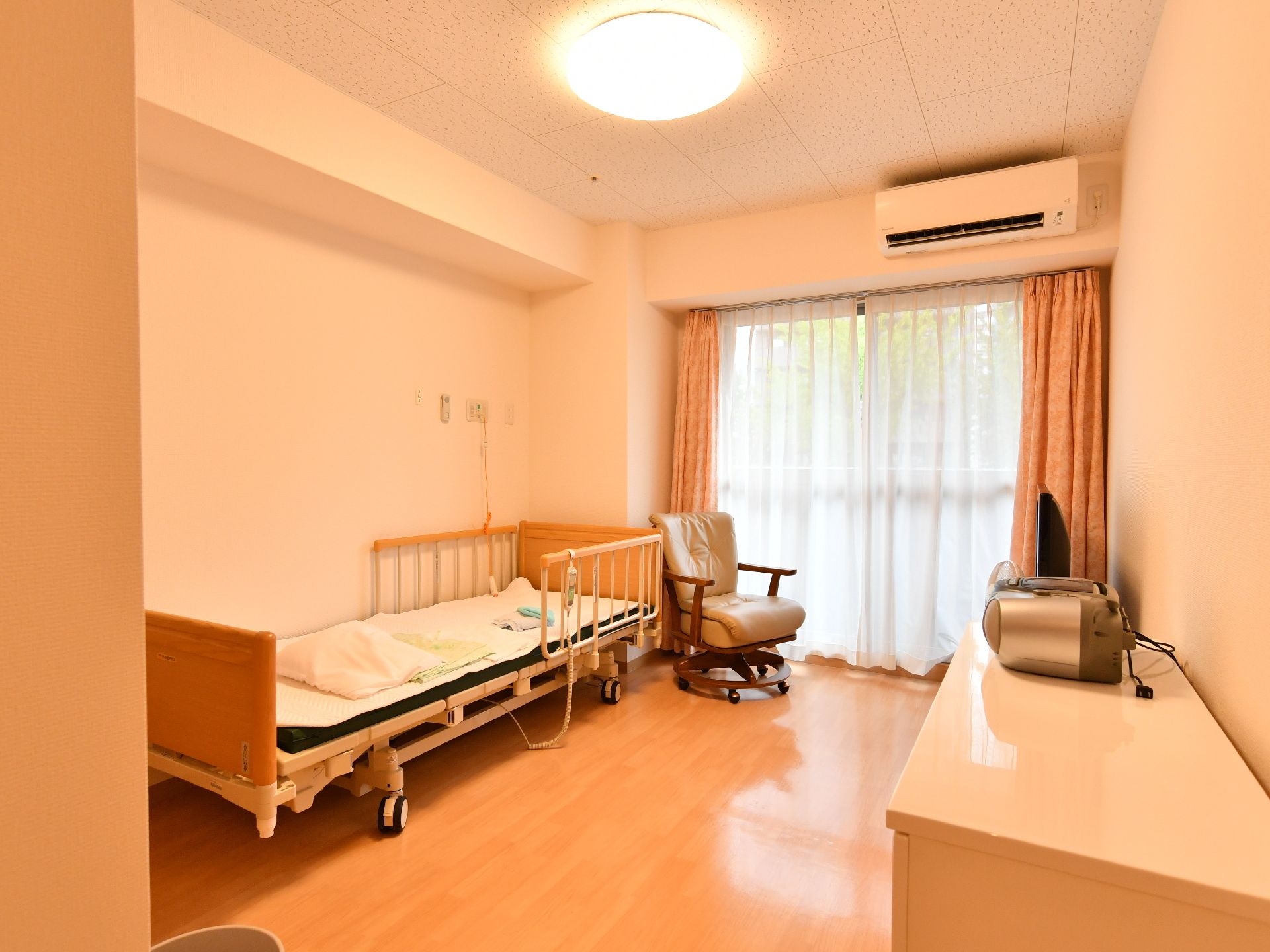 左側には介護用ベッドが置かれている。そのそばには椅子が置かれている。奥には窓が付いており、その上側にはエアコンがある。
