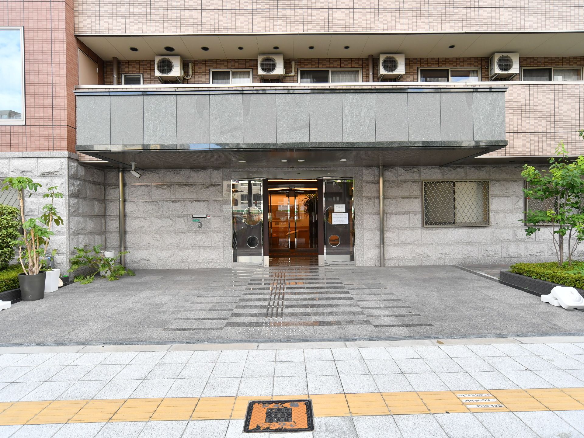 玄関周辺の外壁は他と違って石造りのようになっている。正面奥には自動ドアがある。手前には点字ブロックが敷かれている歩道がある。