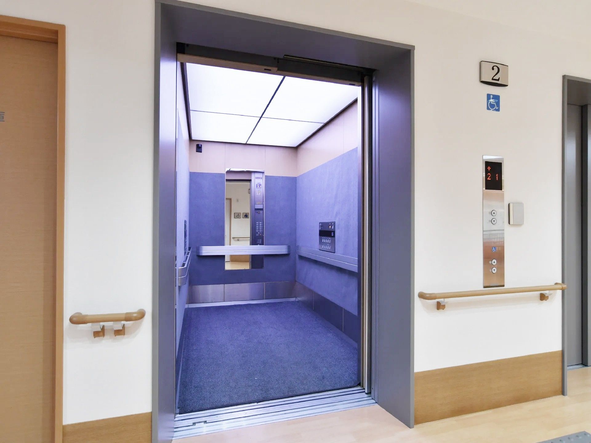 正面にはエレベーターが扉の開いた状態である。その右側には昇降ボタンがあり、車いす用も付いている。周辺の壁には手すりが付いている。