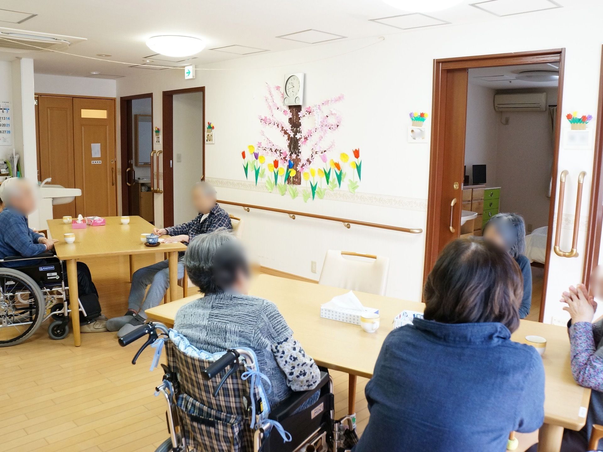 食堂には、長方形のテーブルと椅子が配置され入居者様が着席している。白い壁には、折り紙で作ったチューリップと桜が絵のように飾られている。