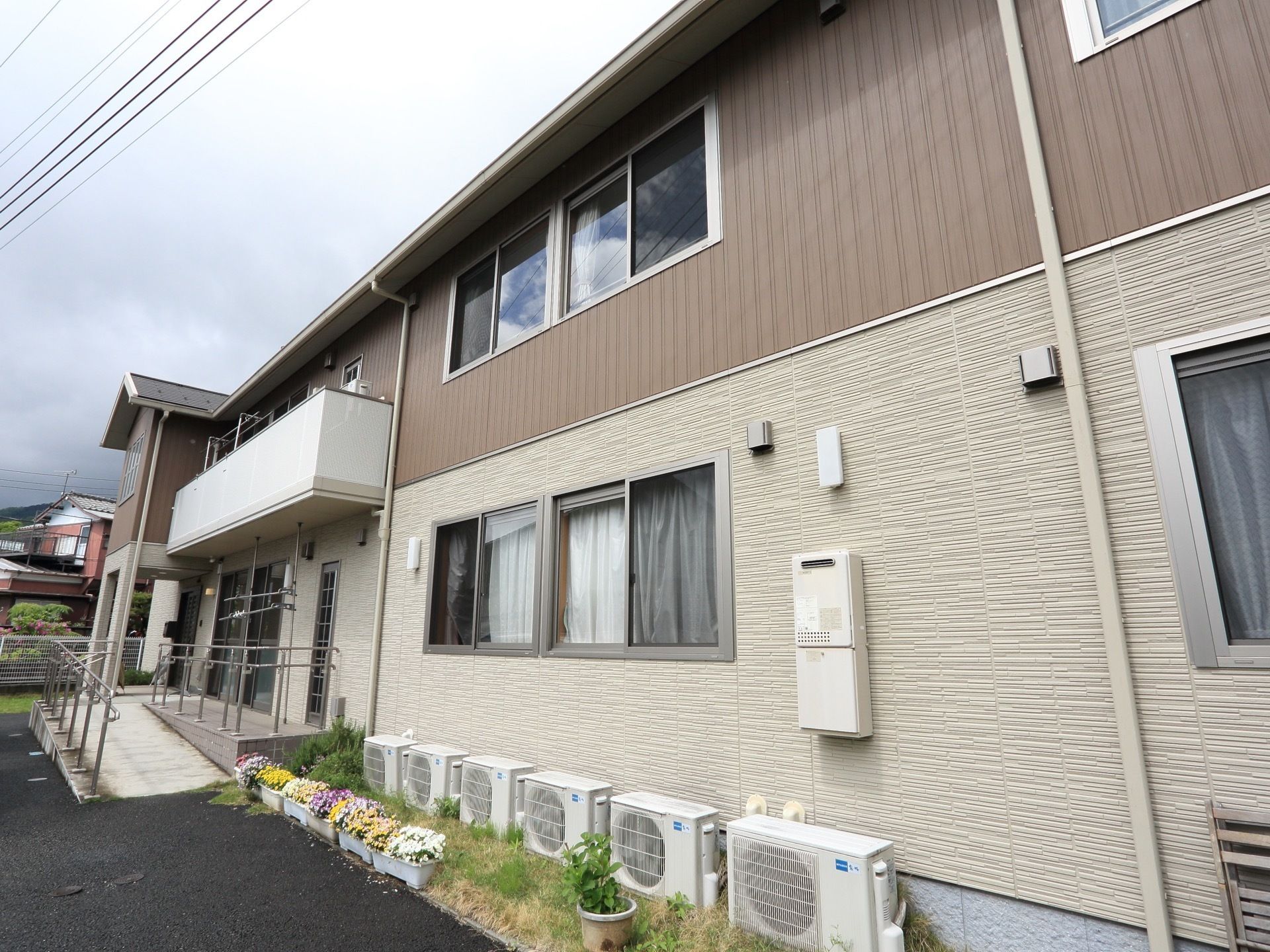 施設の玄関には段差があるので、スロープも設置されている。建物の外壁に沿って置かれているエアコンの室外機の前に、花のプランターが並んでいる。