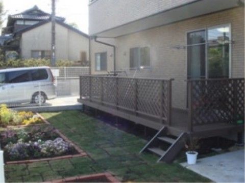 施設の建物の壁はベージュ系で、１階の居室にベランダが設けられている。柵の外には花壇があり、色とりどりの花が咲いている。
