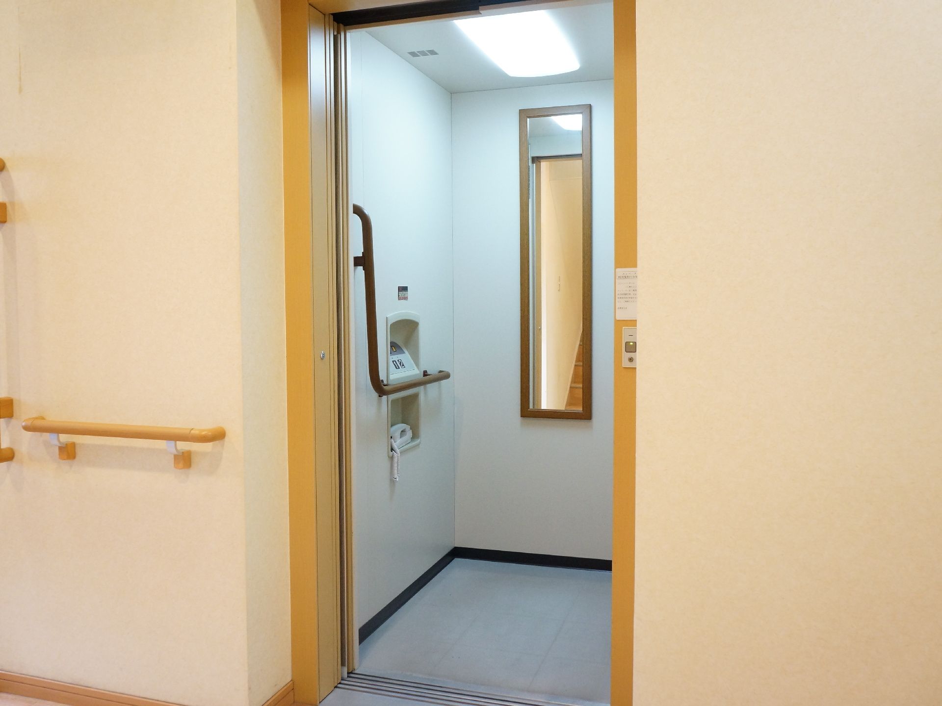 施設内に設置されたエレベーターの扉が開いている写真。内部には非常用の受話器やL字の手すり、奥の壁には鏡が取り付けられている。