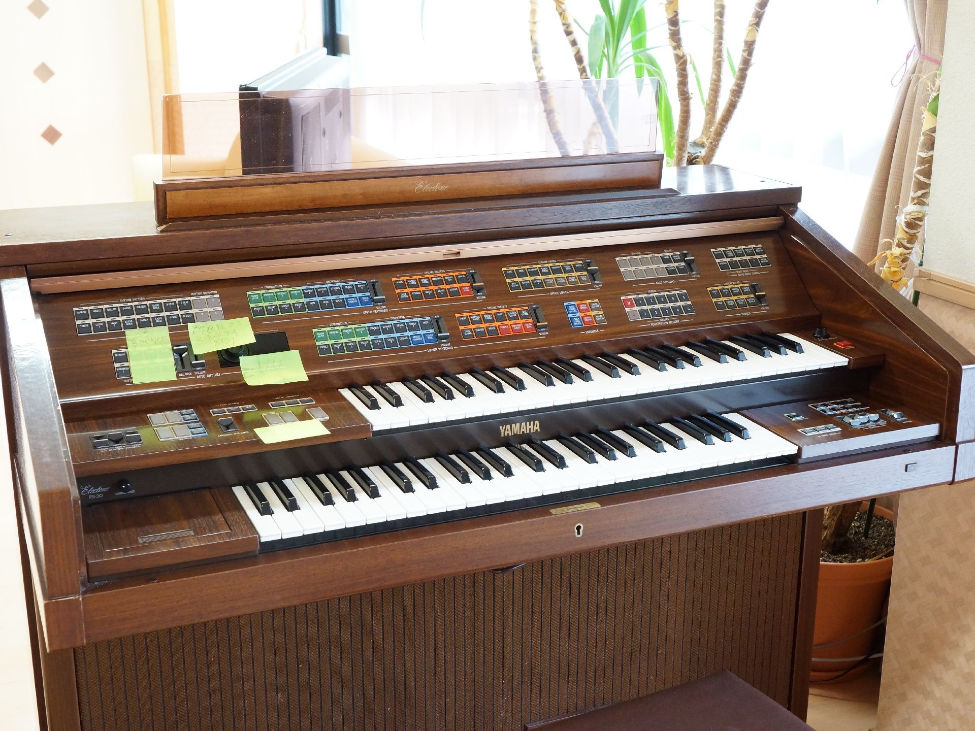 部屋の一角には置かれた電子オルガンの写真。操作用のボタンが沢山ついている。部屋には窓があり、観葉植物が置かれている。