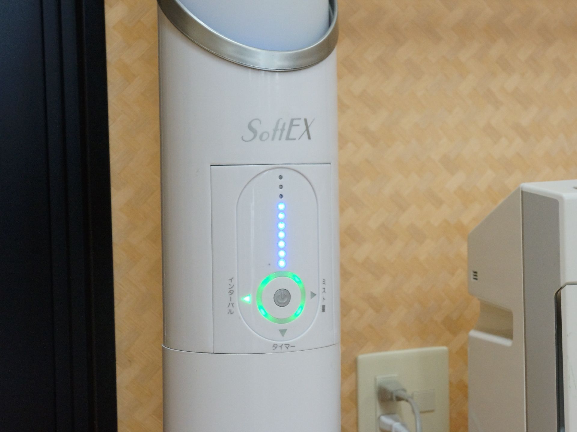 部屋の一角に置かれた次亜塩素酸噴霧器の写真。色は白で細長い形をしている。ボタンでタイマーなどの設定ができる。