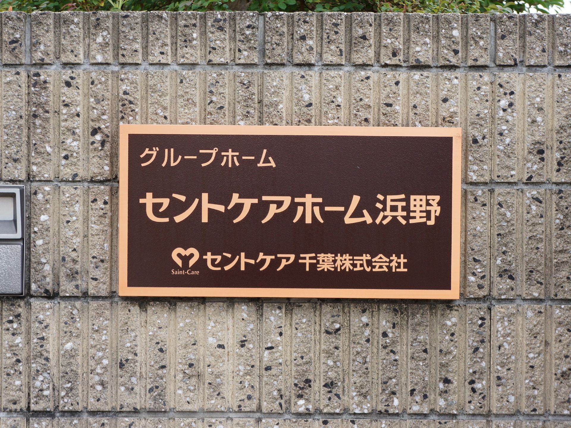 施設の入り口付近にはブロック塀があり、ダークブラウン地にベージュ色で施設名が書かれたプレートが設置されている。