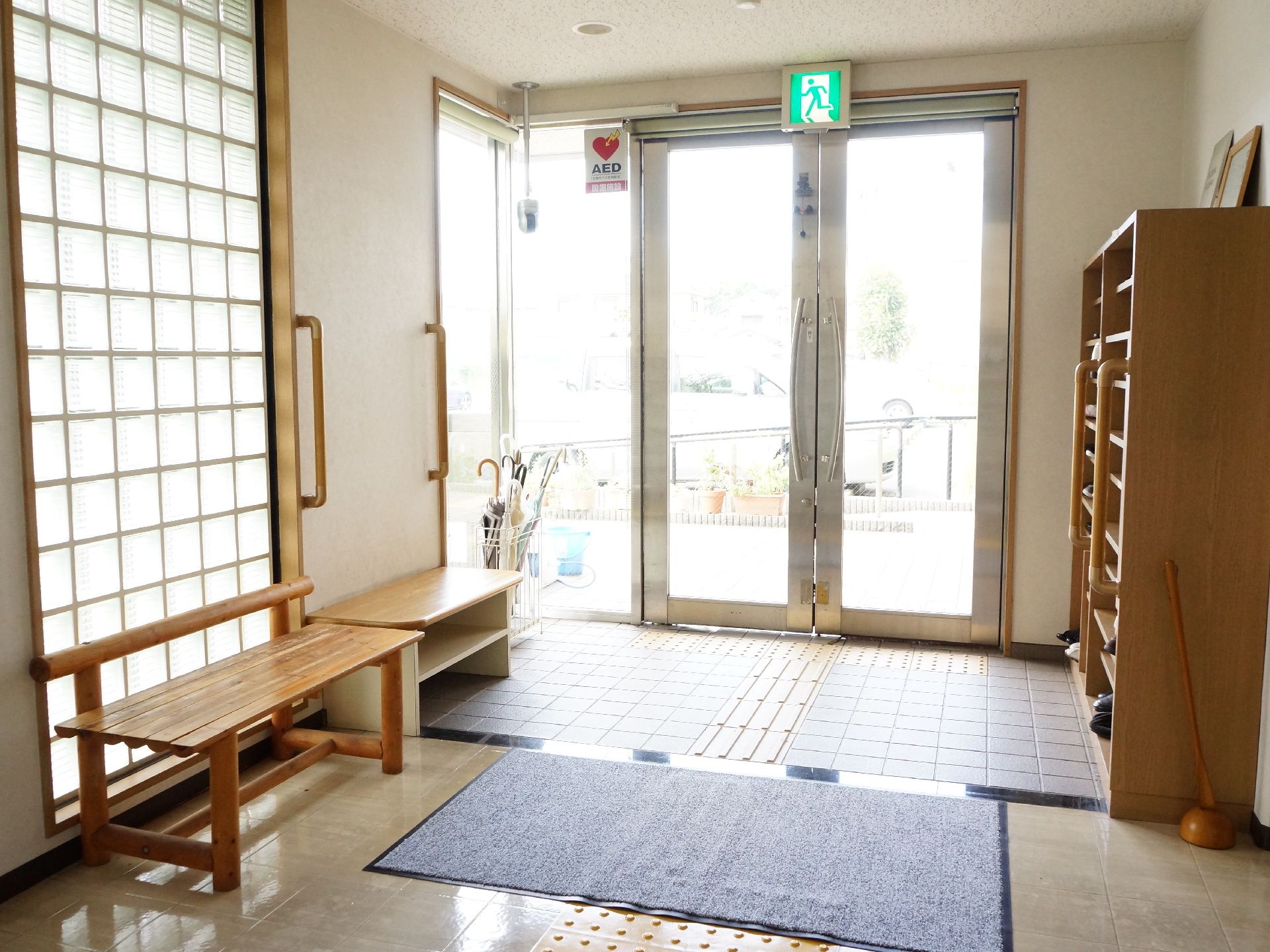 施設の玄関ドアはガラス張りで大きな握りハンドルが付いている。中央部分の天井には非常口の表示灯が設置されている。