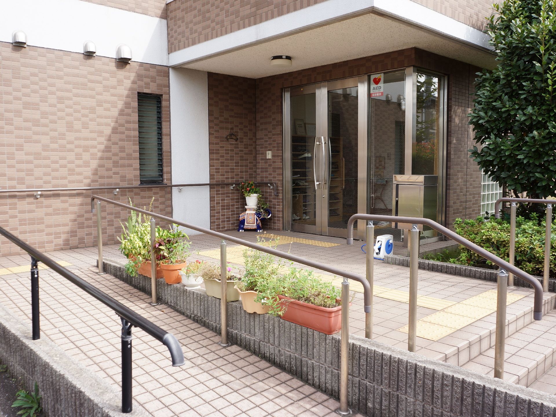 施設の玄関周りはガラス張りで、上部にはAEDの大きなシールが貼られている。ドア前には黄色い点字ブロックが設置されている。