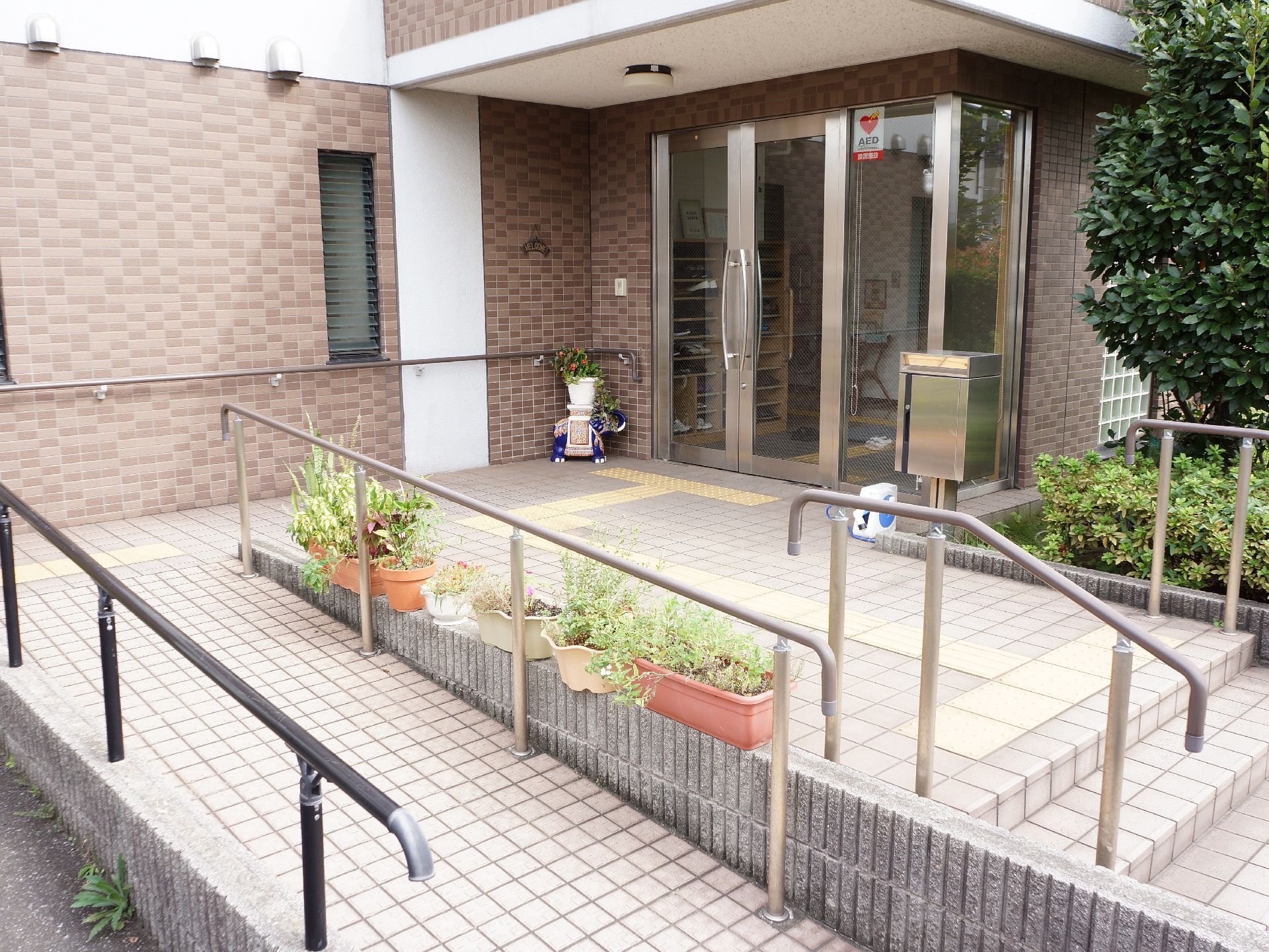 施設の入り口付近の床はブラウン系のタイル敷きある。階段とスロープがあり、間を仕切るブロックの上には植物の鉢が置かれている。
