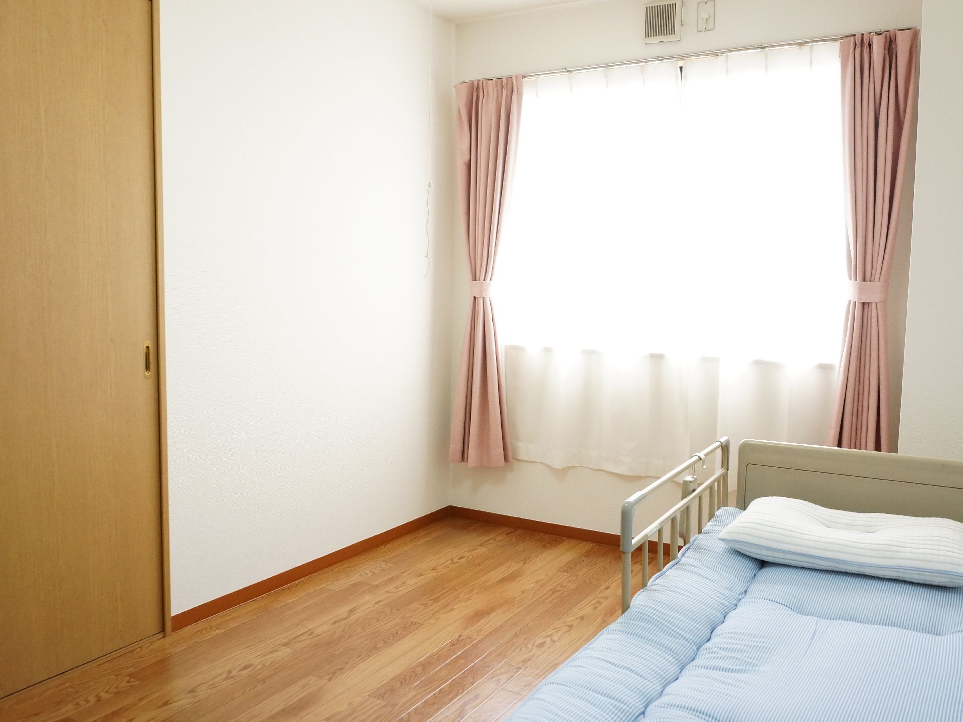 居室の1方向の壁には腰高窓があり、白いレースとピンクの厚地のカーテンが掛けられている。右側にベッドが置かれている。