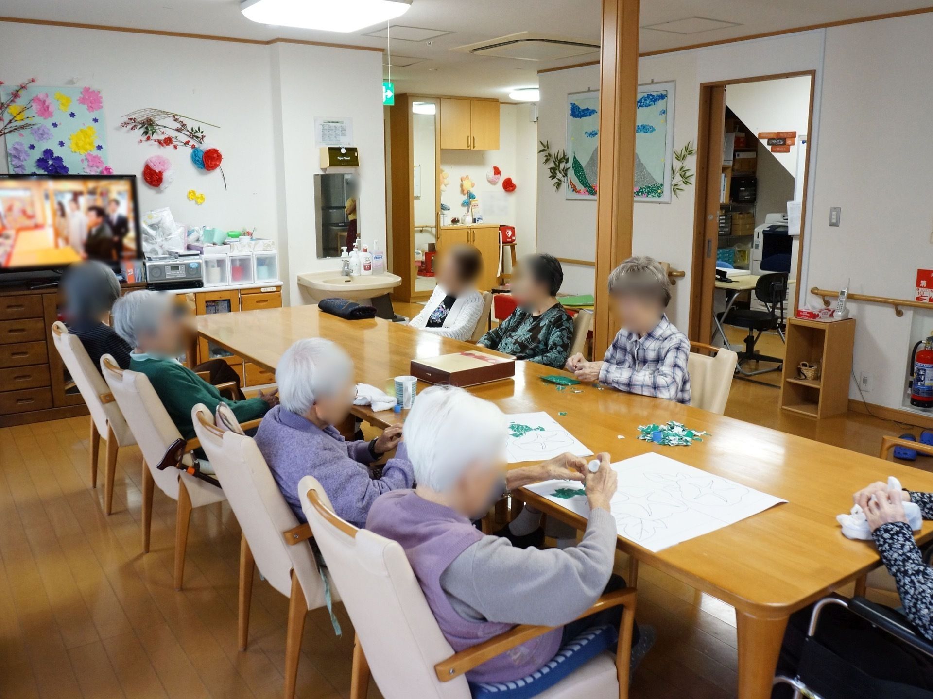 長テーブルが用意されており、入居者様が座っている。ちぎり絵を作成している最中である。壁際にはテレビが置かれている。