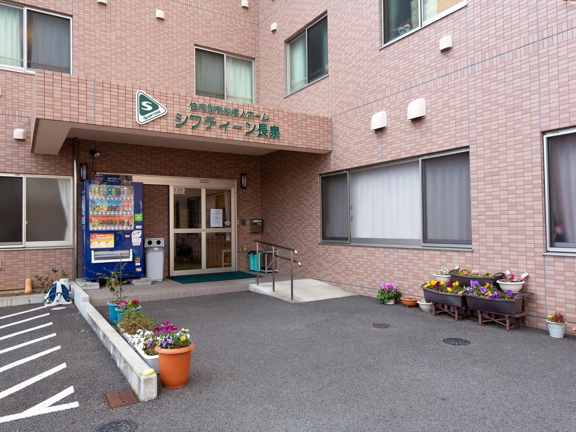玄関にはひさしがついている。ひさしの前面に緑色の文字で施設名称が表示されている。玄関に飲料の自動販売機がある。