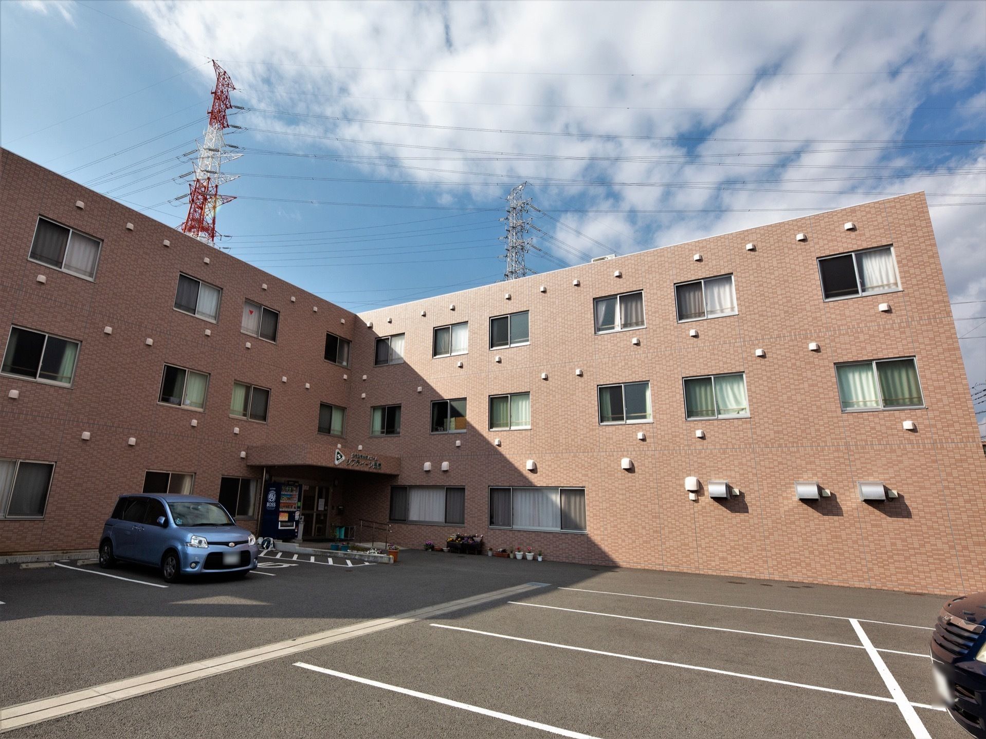 くの字型になっている３階建ての施設である。コーナー部に玄関がある。敷地内に駐車スペースがたくさんある。