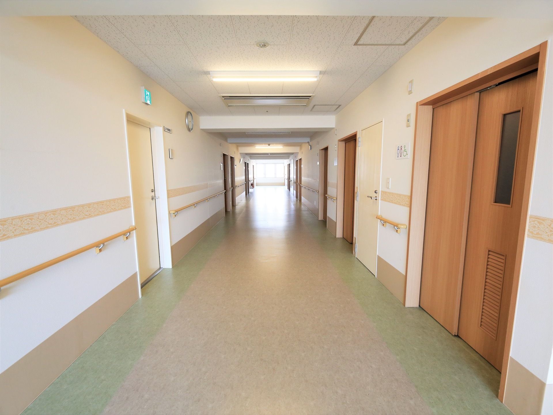 つきあたりの大きな窓からの採光や照明で明るい廊下である。白い壁と居室入口の色の組み合わせが、落ち着いた印象を与えている。