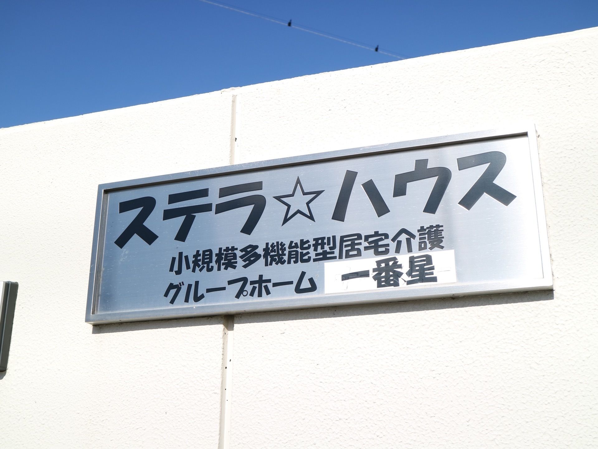 白を基調とした凹凸のある外壁に、看板が取り付けられている。銀色の四角いパネルに、黒い文字で施設名が書かれている。