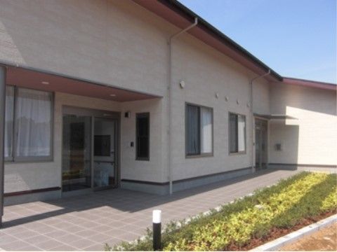 茶系の落ち着いた雰囲気の施設の外観である。平屋のように横長の造りで、エントランス前の花壇には植物が植えられている。