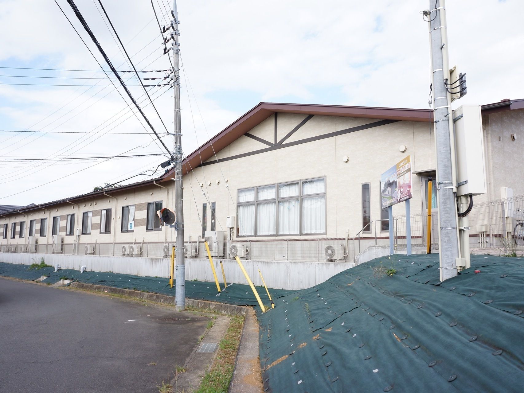 正面から左奥にかけて建物が建っている。正面方向には窓が複数付いており、その手前には室外機が置かれている。左側には道路がある。