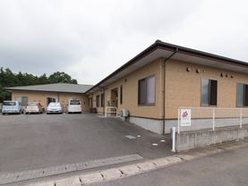 広々とした車の出入りにも充分な入口スペースの奥に建物と隣接して駐車場が設けられている。建物は平屋建てのシンプルで親しみのあるデザインである。