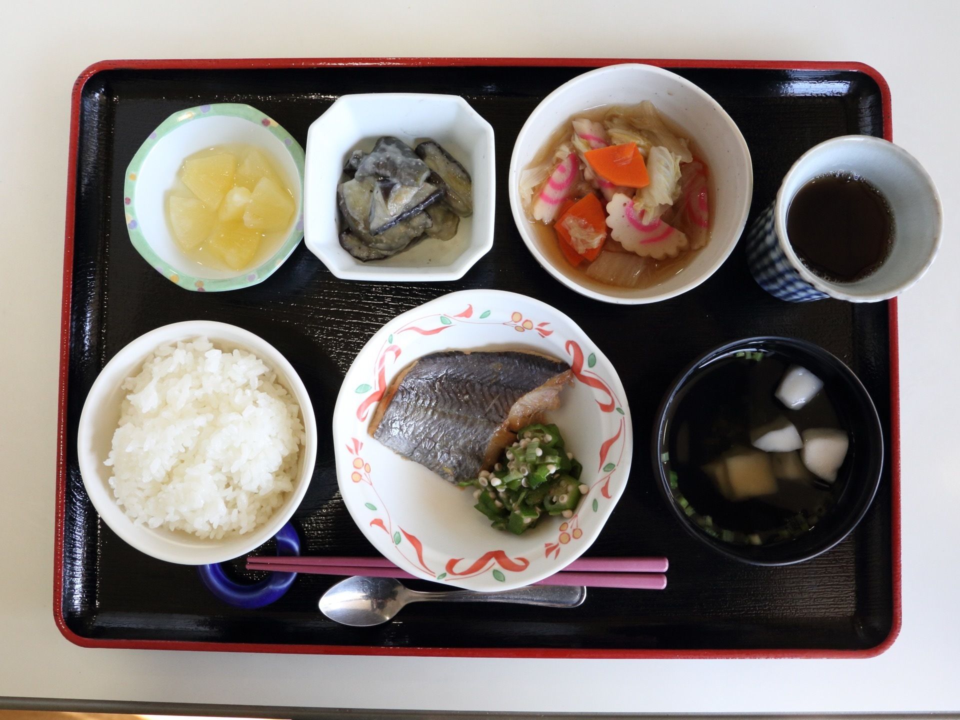 黒いお盆の上にはご飯、魚、汁椀、なるとと野菜の煮物、和え物、フルーツ、お茶が置かれている。お箸とスプーンが添えられている。