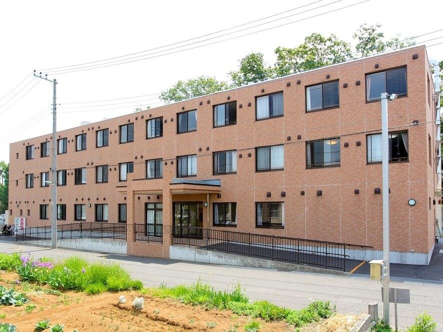３階建ての大きな施設は、遠くからでも見付けやすい。周囲は住宅街という落ち着いた環境である。入居者様募集の案内が掲げられている。