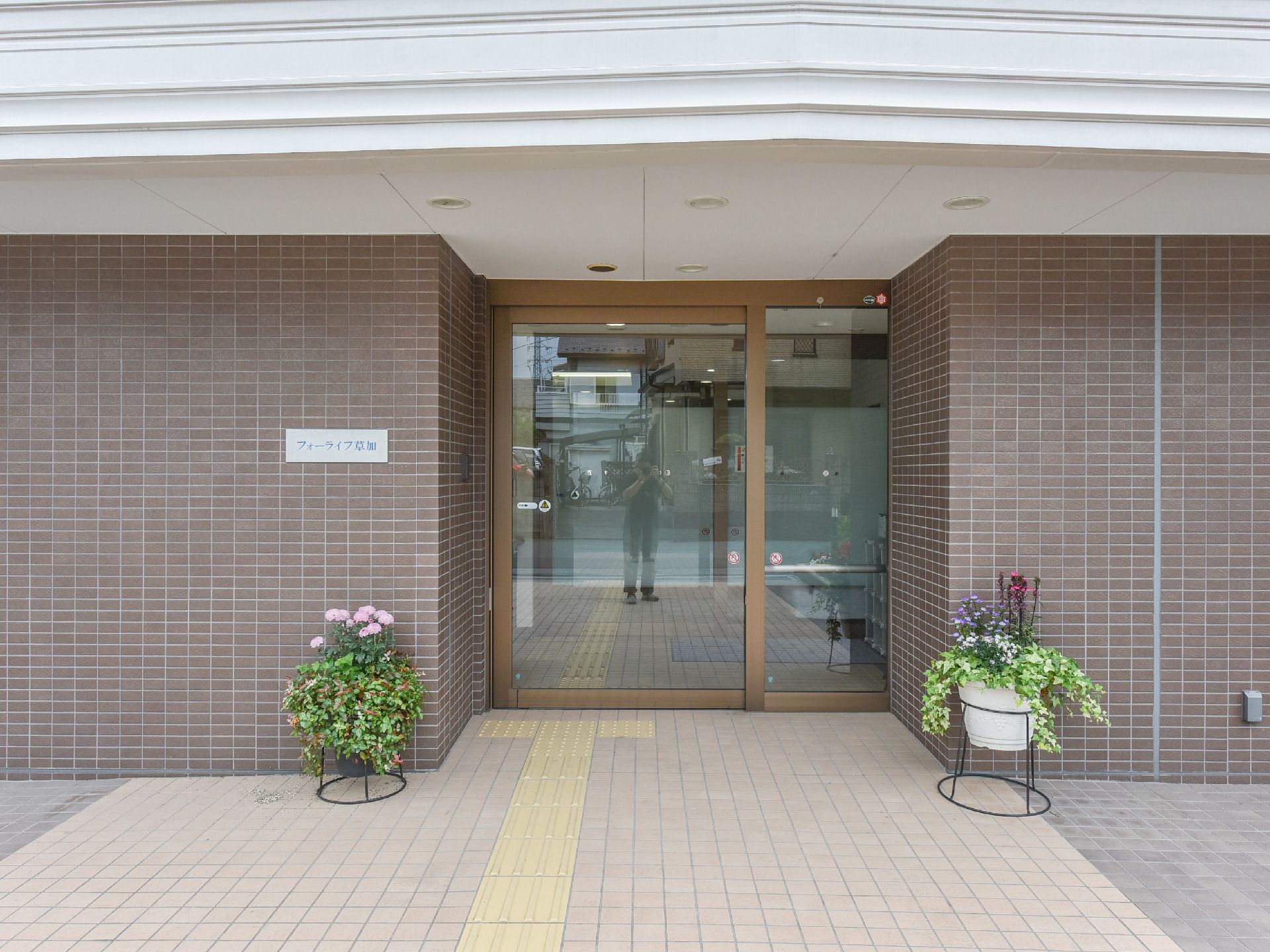 エントランスには、目の不自由な方に配慮して入口の自動ドアまで誘導ブロックが設置されている。花のプランターが両サイドの壁際に置かれている。