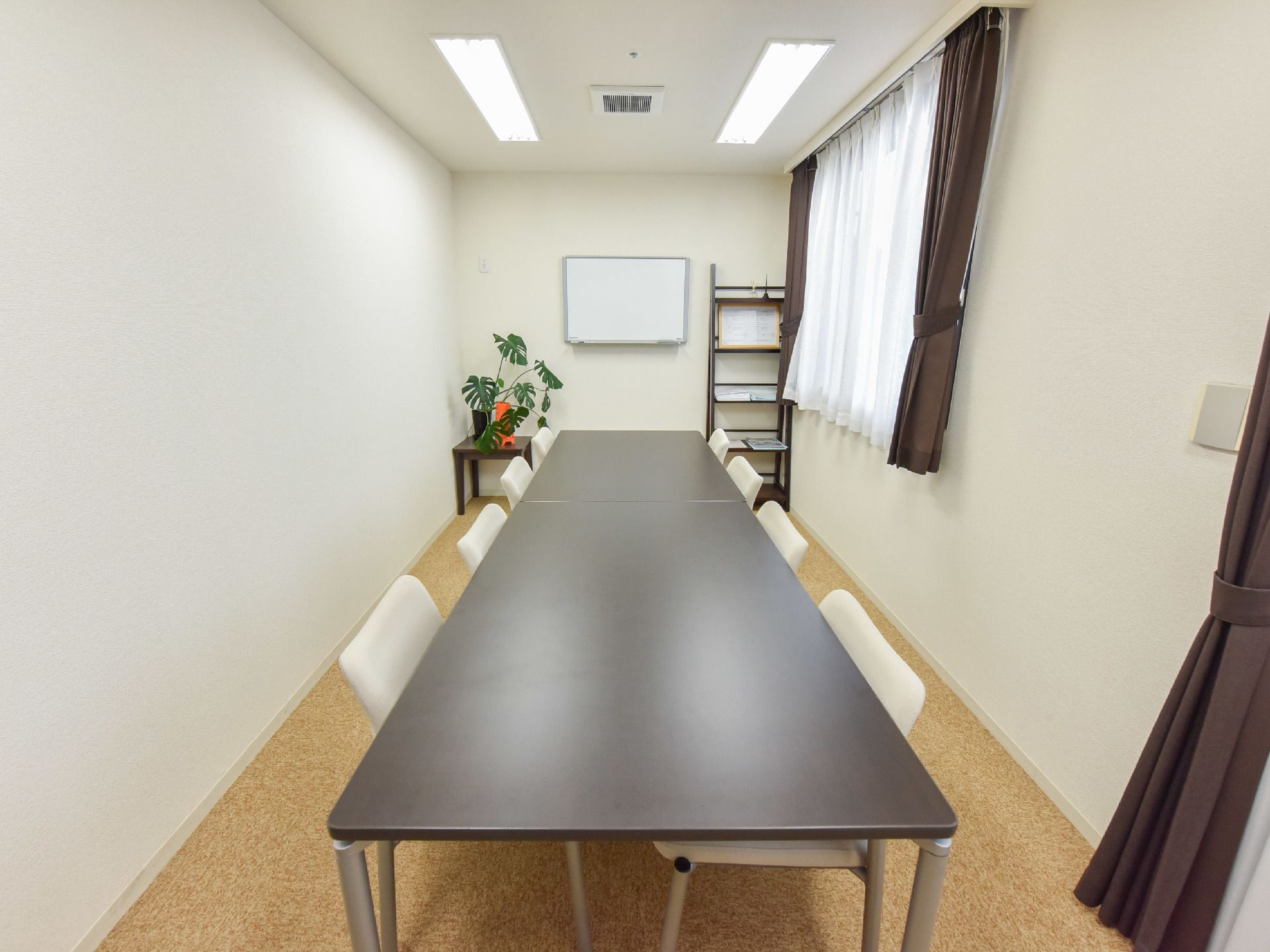 部屋の奥には、観葉植物が置かれた台と棚が置かれており、ホワイトボードが壁に取り付けられている。縦に4人掛けのテーブルが2台配置されている。