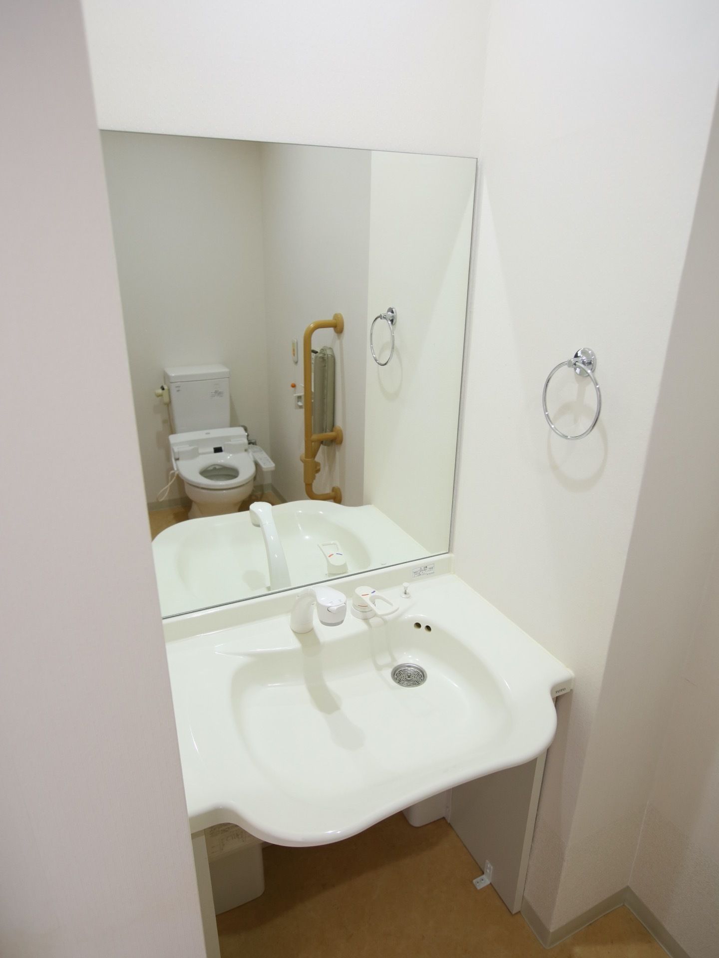 トイレには洗面台が備え付けられている。壁には手すり、ペーパーホルダーが取り付けられている。壁は白である。