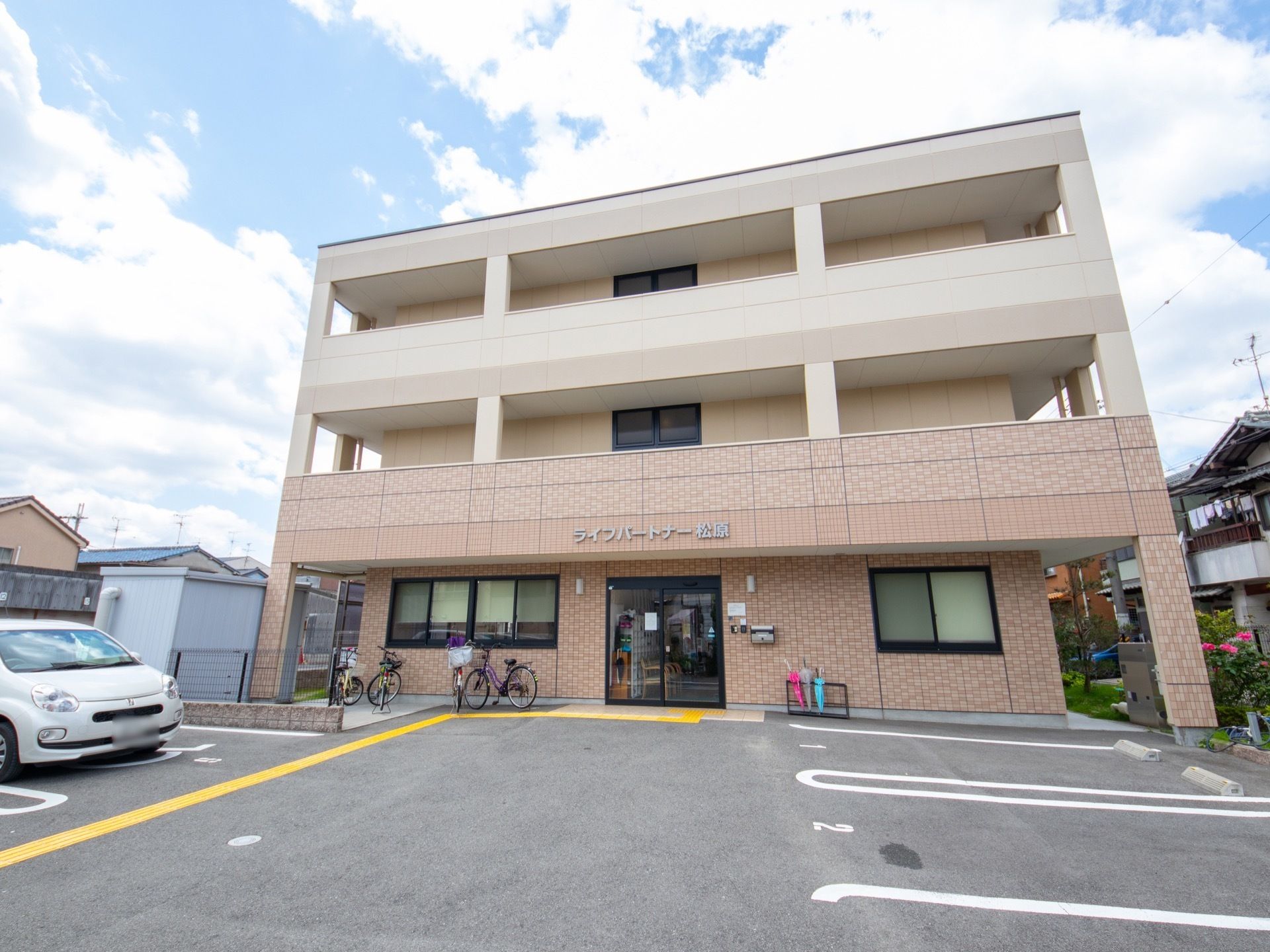 一般的なアパートのような外観で駐車場がある。自転車が停まっている。入口横には傘立てが設置されている。