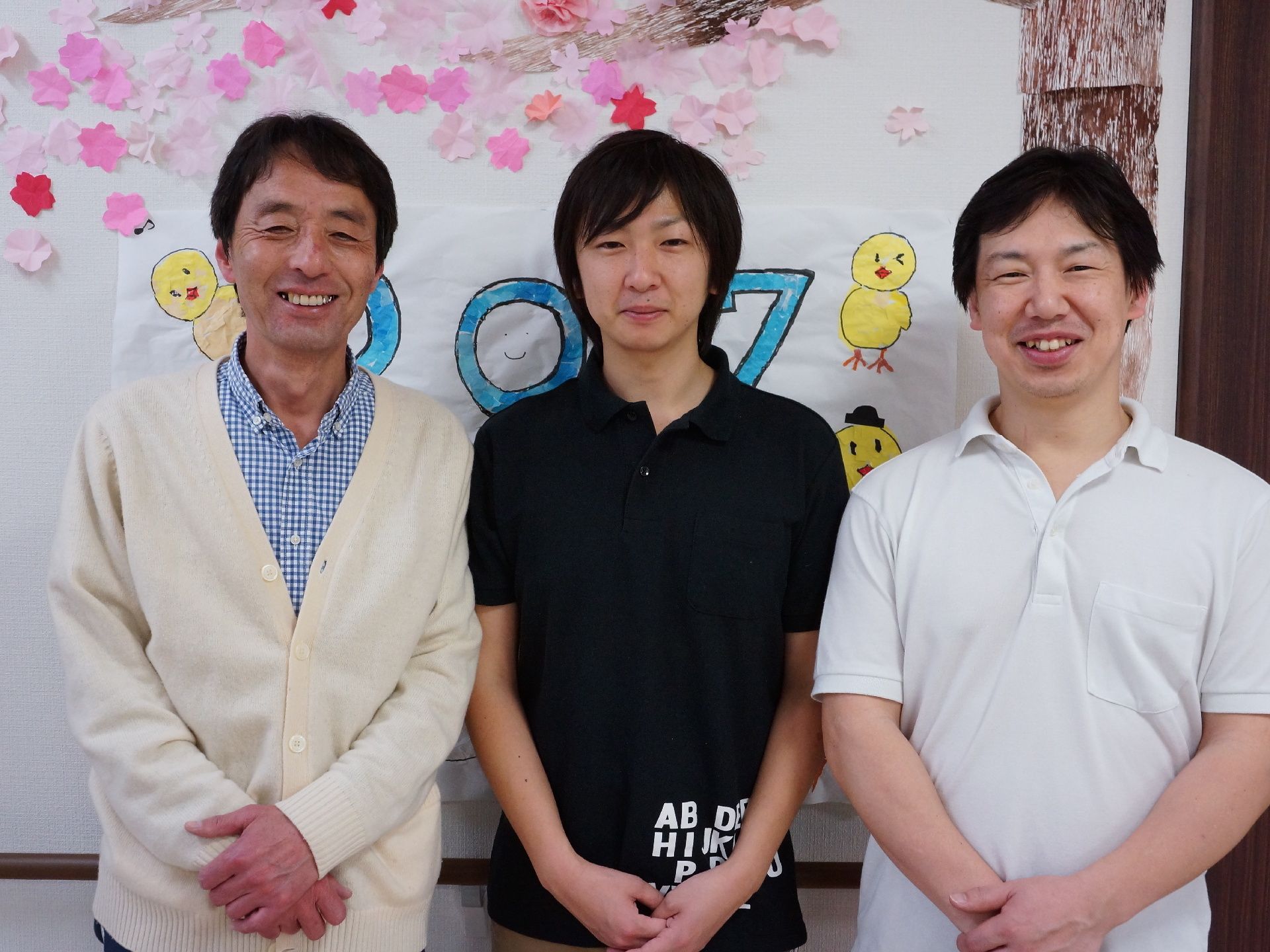手作りの飾りつけがされた壁の前に立つ、笑顔の男性スタッフ