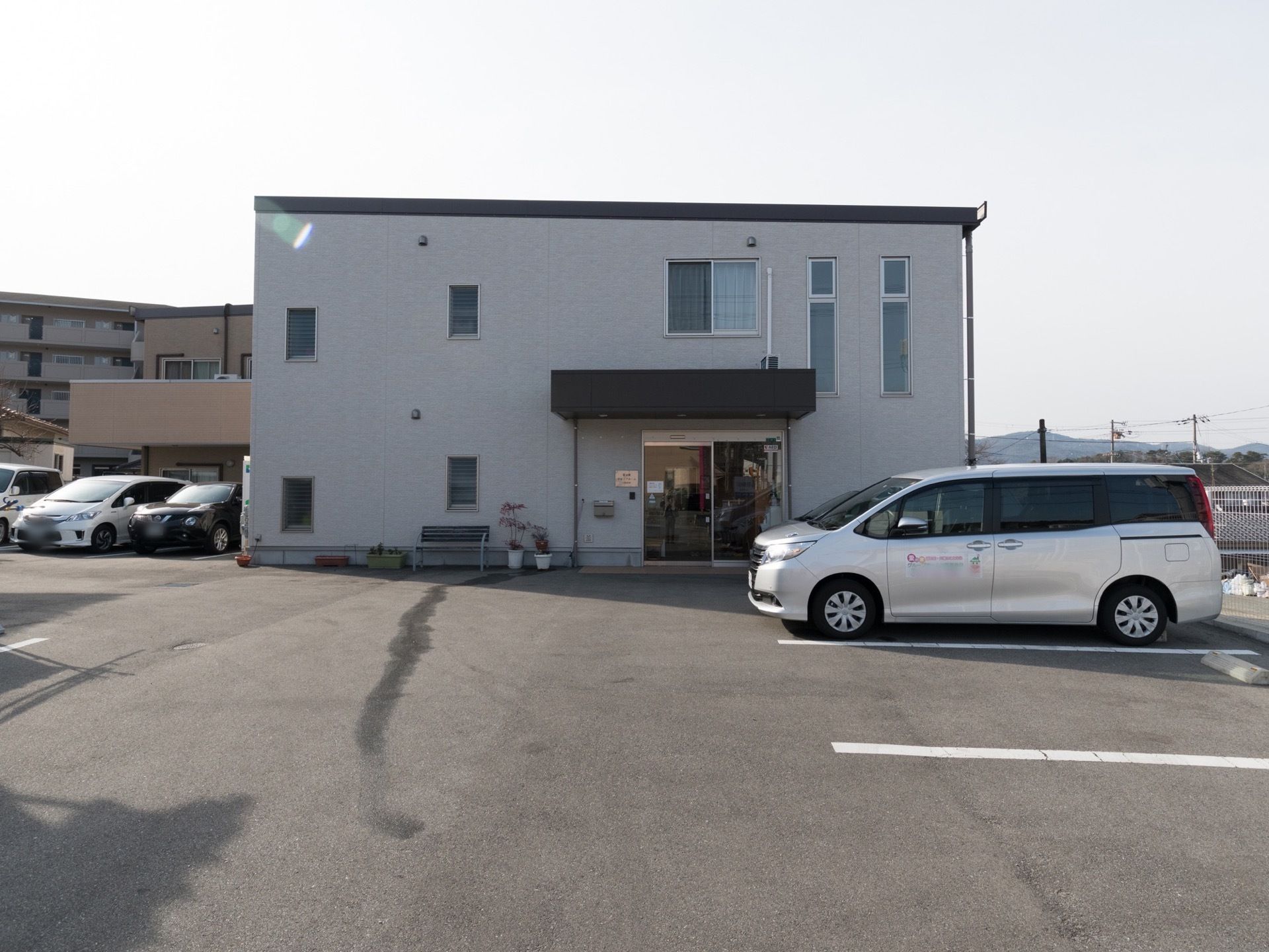 白色の外壁で、２階建ての建物である。自動ドアが設置されている。施設車両が玄関のすぐ前に駐車されている。