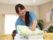 女性スタッフがタオルをたたんで運んでいる。黄色のソファーが並べられており、観葉植物も飾られている部屋である。