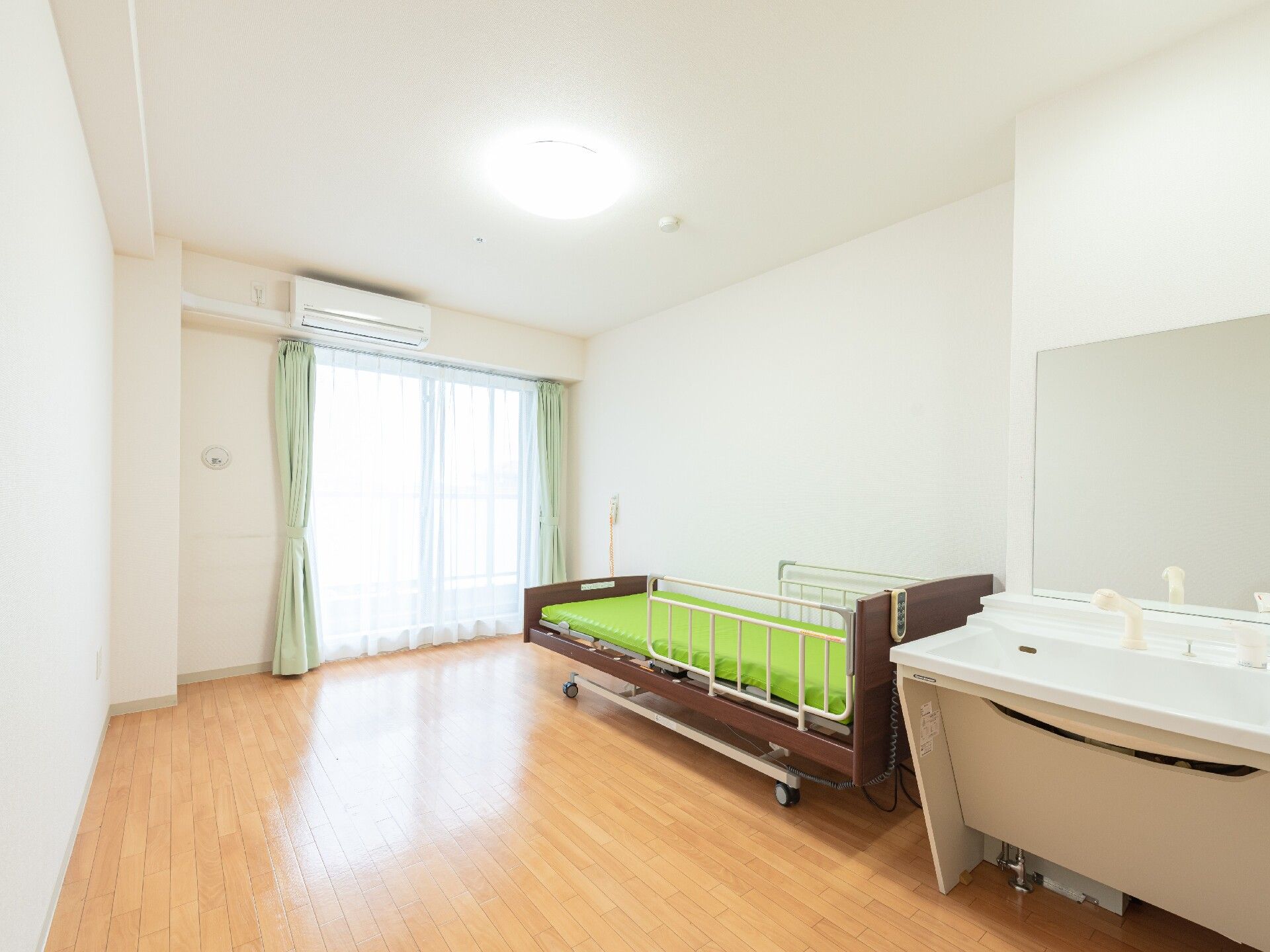 木目調の床に白い壁や大きめの窓がある明るい居室である。壁にはナースコールが付いており緊急時などには安心である。